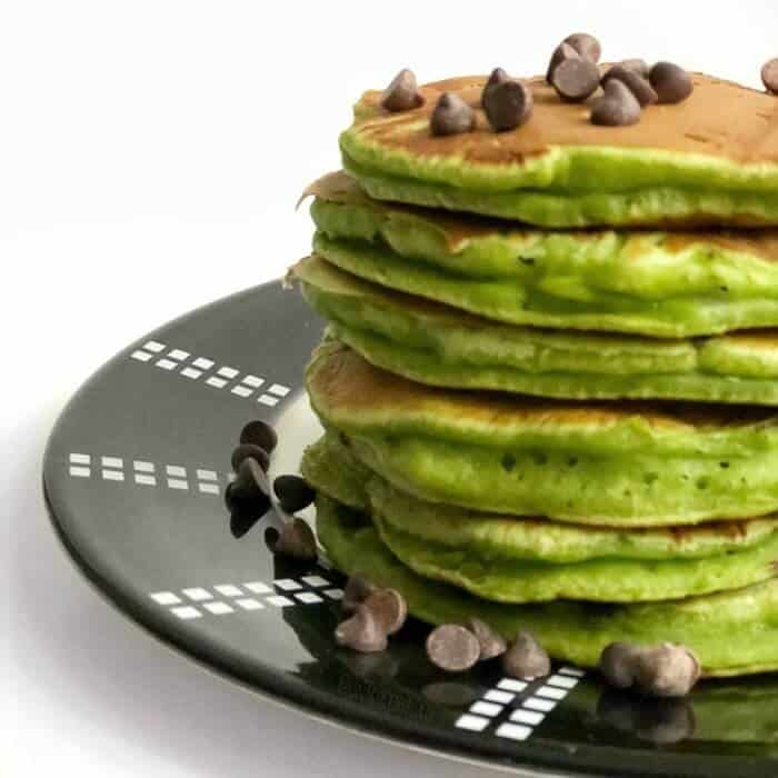 A stack of green spinach pancakes sits on a plate with a sprinkle of chocolate chips on top.