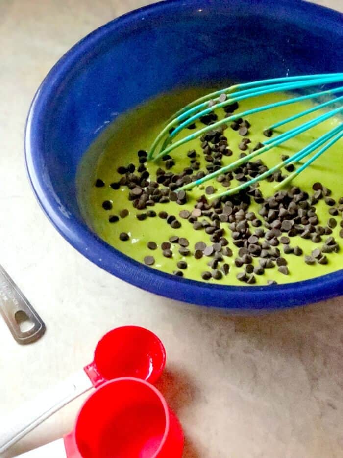 Green spinach pancake batter sits in a bowl with a whisk and measuring spoons. A sprinkle of mini mint chocolate chips is on top of the batter.