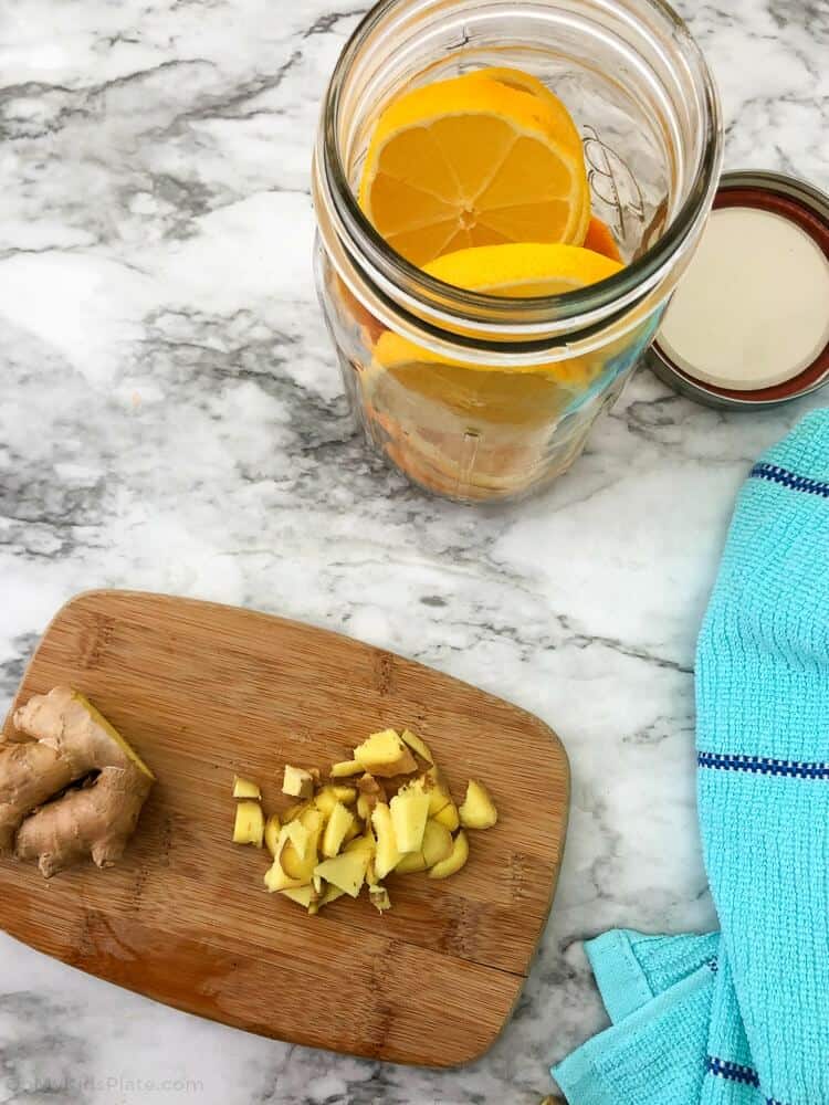 Ginger being diced on a cutting board from above next to a mason jar full of sliced oranges
