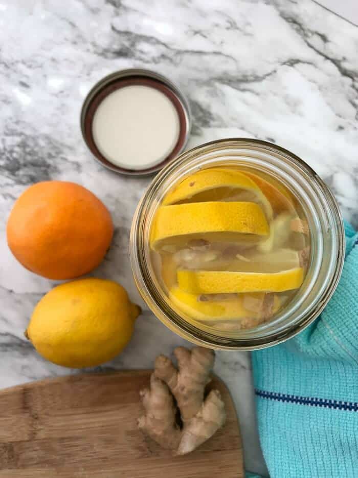A mason jar from overhead full of sliced lemons next to a lemon, orange and ginger on a cutting board