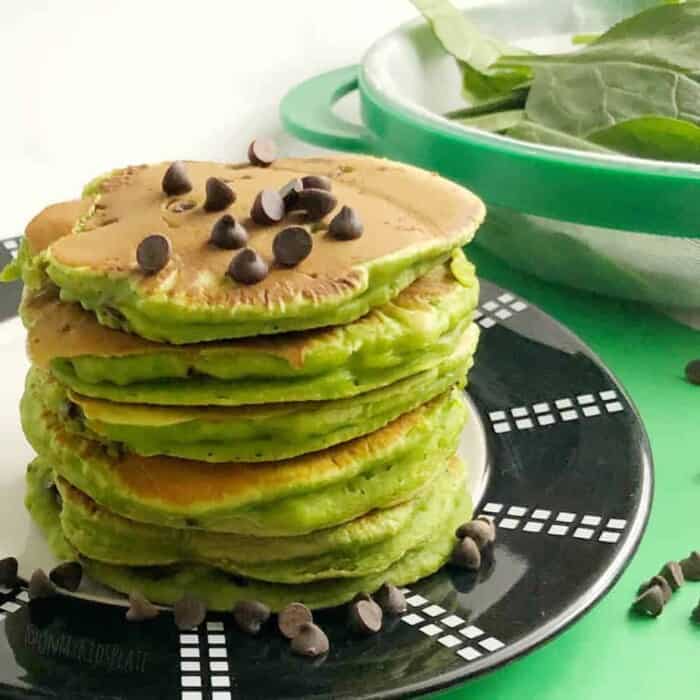 A stack of green spinach pancakes sits on a plate with a sprinkle of chocolate chips on top.