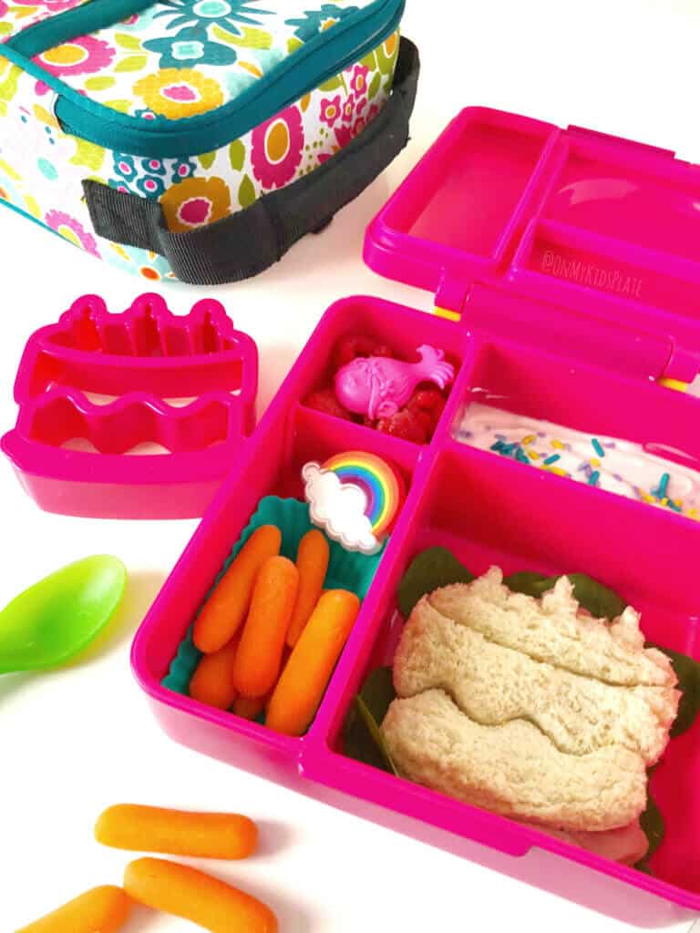 A child\'s bento lunchbox full of carrots, yogurt topped with sprinkles and a sandwich shaped like a birthday cake.