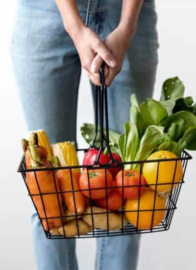 A mom stands holding a grocery basket full of fruits and vegetables, we only see her legs and the basket.
