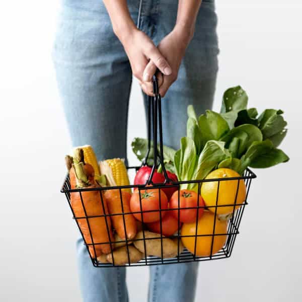 A mom stands holding a grocery basket full of fruits and vegetables, we only see her legs and the basket.