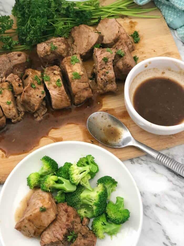 A plate of pork tenderloin and broccoli from overhead next to a serving platter, bowl of sauce and spoon.