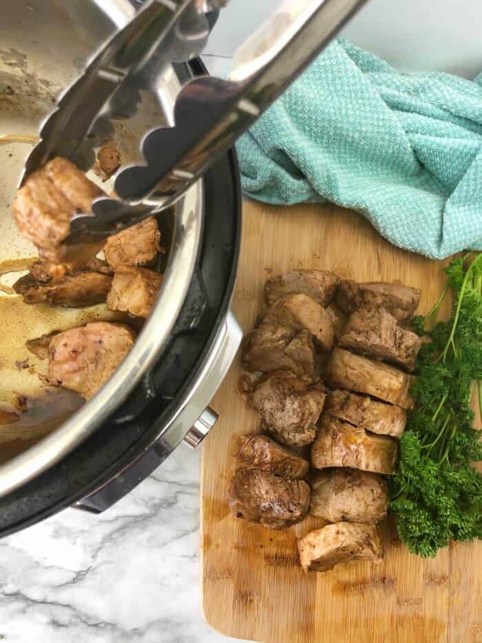 Pork tenderloin pieces being removed with tongs from an instant pot from overhead to a cutting board full of sliced pork pieces.
