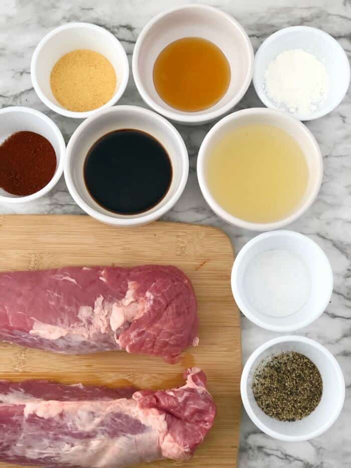 Two raw pork tenderloins on a cutting board surrounded by small bowls of spices and sauce ingredients.
