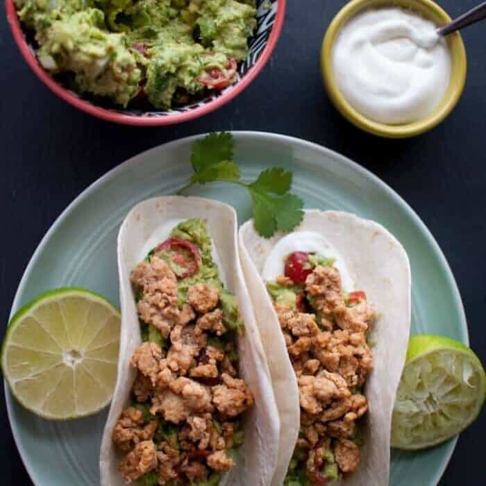 A plate holding two tacos and lime with guacamole and sour cream in bowls next to the plate.