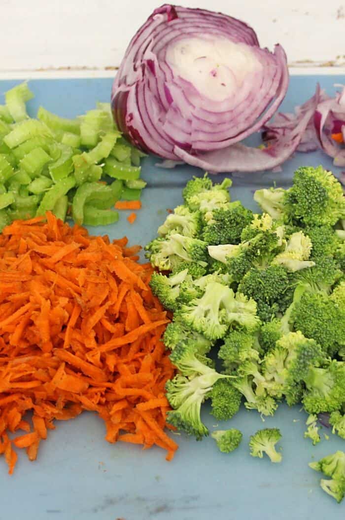 Carrots, celery purple onion and broccoli all sit on a cutting board chopped and ready to add to the broccoli apple salad with pasta bowl.