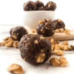 A bowl full of chocolate walnut energy bites sits on a counter with a few snack bites front and center surrounded by fresh walnuts.