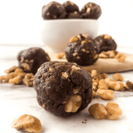 A bowl full of chocolate walnut energy bites sits on a counter with a few snack bites front and center surrounded by fresh walnuts.