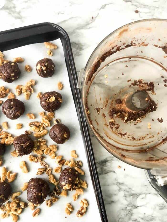 Energy bites rolled on a baking sheet with walnuts scattered around next to an empty food processor.