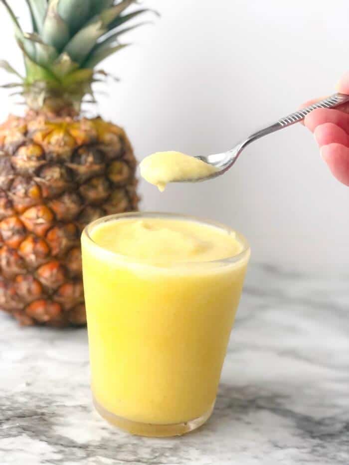 A pineapple dole whip smoothie being lifted over a glass with a spoon, a pineapple in the background.