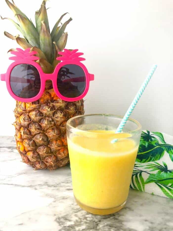 A glass of yellow smoothie with a straw next to a pineapple wearing kid\'s pineapple sunglasses.