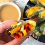 A hand holds a mango chicken fresh spring roll and we can see the veggies and meat inside. Below on the counter is a bowl of peanut dipping sauce and a platter for fresh spring rolls