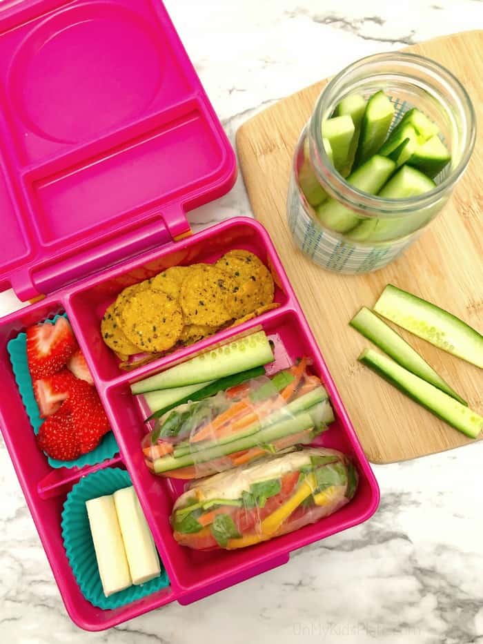 A kid\'s bento lunchbox filled with fresh spring rolls, cucumber, sweet potato crackers, strawberries and sting cheese. A jar full of cucumber slices sits on a cutting board next to the lunchbox.