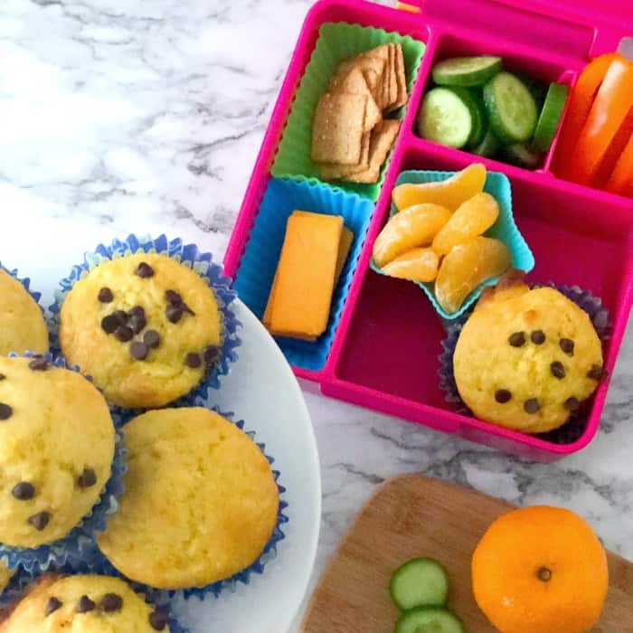 A kid\'s bento lunchbox with an orange muffin, oranges, pepper, cucumber, crackers and cheese next to a platter of muffins