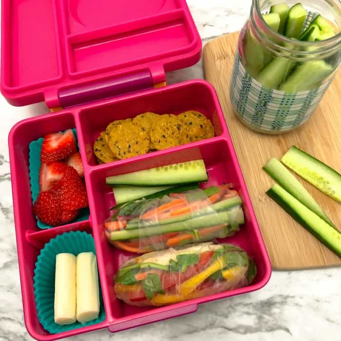 A kid\'s bento lunchbox filled with fresh spring rolls, strawberries, cucumber sticks, sweet potato crackers and string cheese