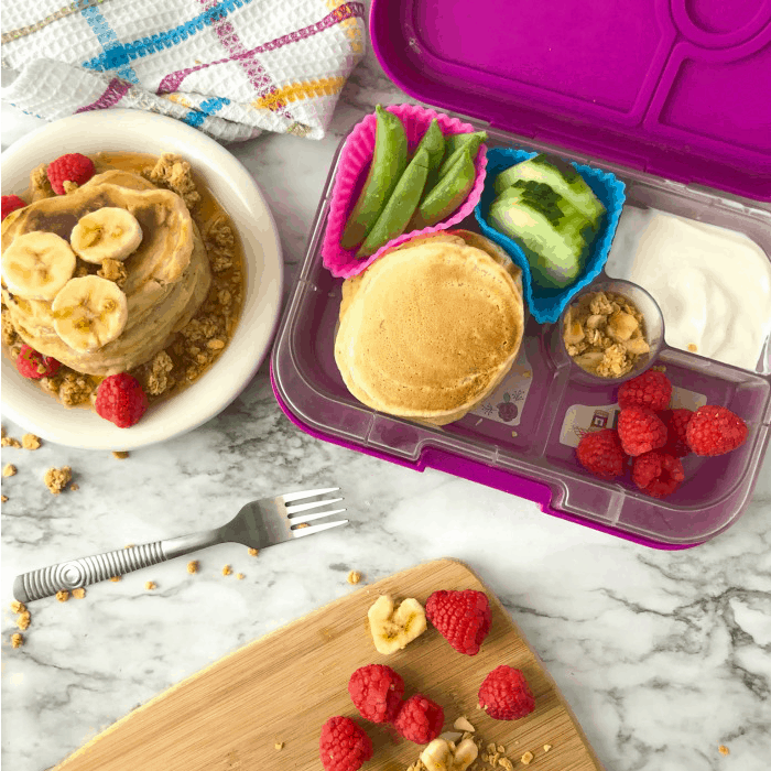 A kid\'s bento lunchbox filled with pancakes, raspberries, yogurt, cucumbers, snap peas next to a stack of pancakes on a plate