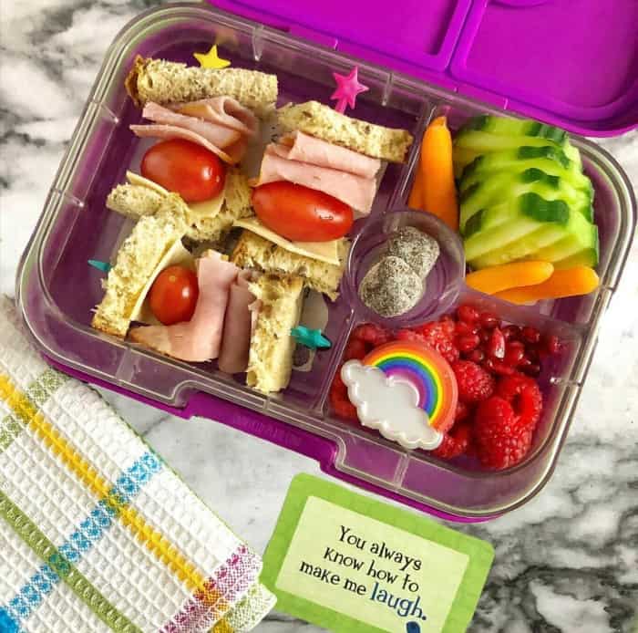 A kid\'s bento lunchbox full of a ham sandwich and cherry tomatoes cut into bites on skewers, cucumbers, raspberries, pomegranate seeds, carrots and  a rainbow.