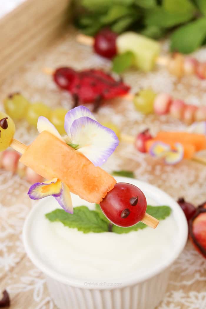 A fruit skewer decorated like a winged insect about to be dipped in a fruit dip