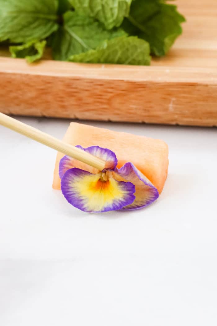 A piece of cantaloupe being pushed with a skewer and an edible flower to make an insect wing.