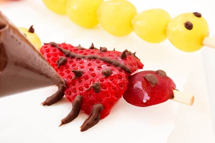 A ladybug made out of a strawberry and grape being decorated with chocolate insect legs