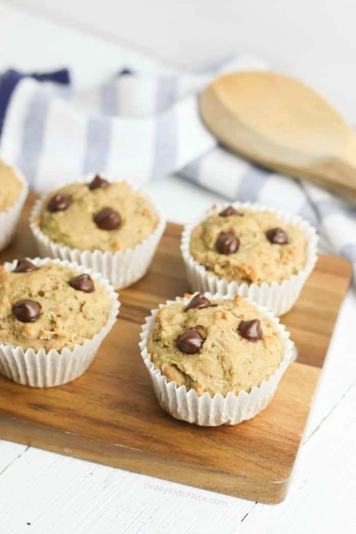 Four muffins with chocolate chips on a cutting board, a wooden spoon and towel behind