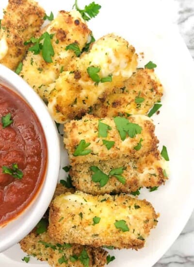 Mozzarella cheese sticks on a plate overhead next to a bowl of sauce topped with parsley