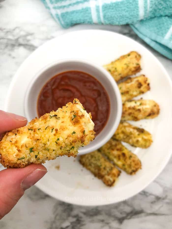 A mozzarella cheese stick being held by fingers over a bowl of sauce and platter of snacks