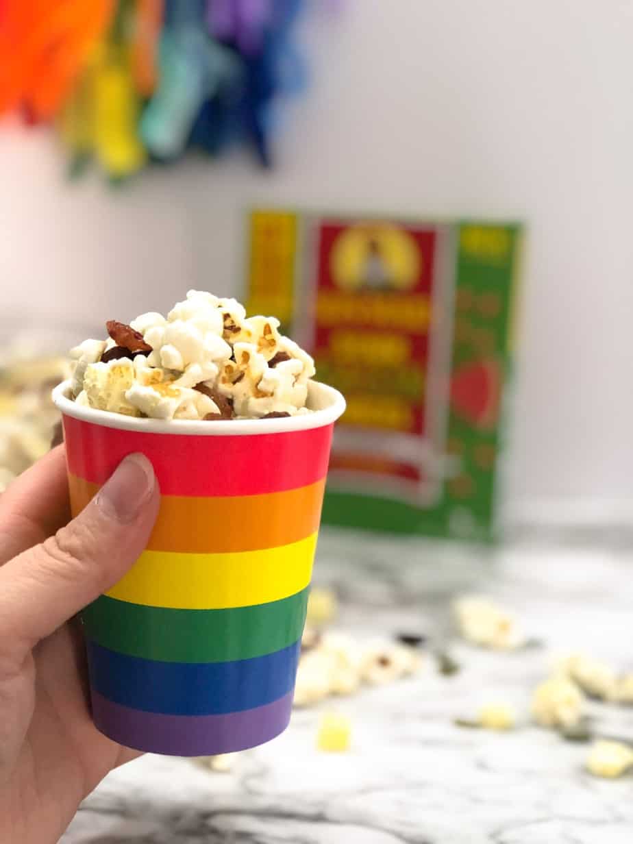 A rainbow colored cup full of popcorn trail mix with a box of raisins in the background