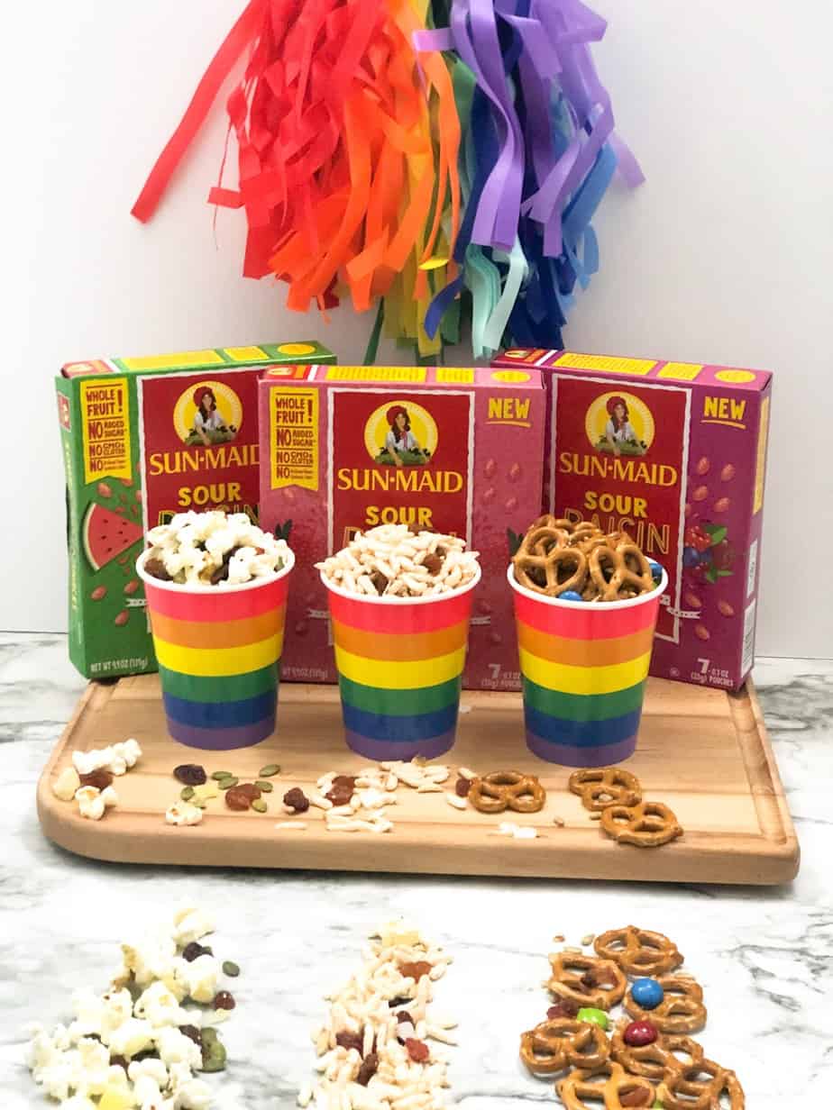 Three different rainbow cups of different trail mixes on a cutting board next to three boxes of flavored raisins