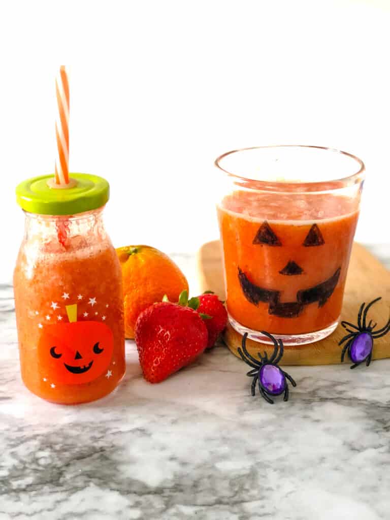 orange smoothie in a cup and milk glass decorated like pumpkins next to oranges and strawberries and spider rings
