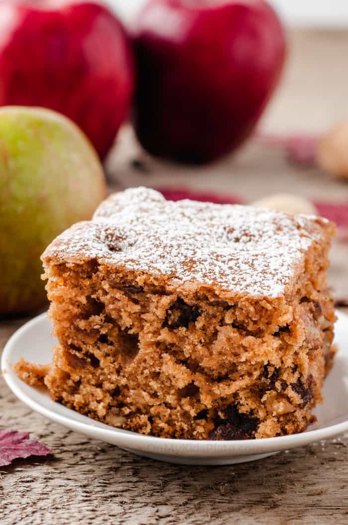 A slice of apple walnut cake sits on a plate with apples in the background.