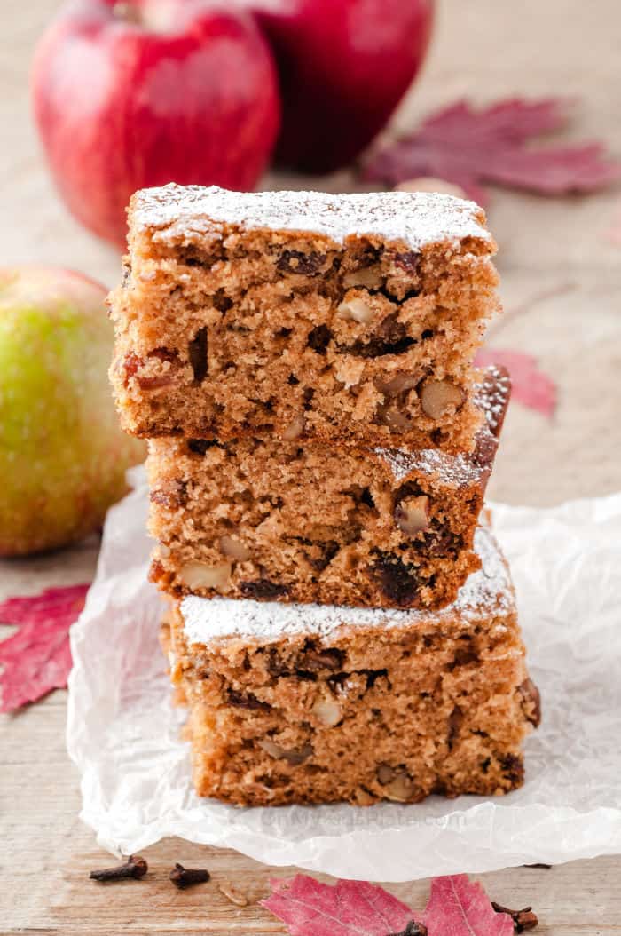 Three slices of cake stacked on top of each other on top of a piece of parchment paper with apples in the background