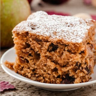 A slice of apple walnut cake sits on a plate with an apple in the background.