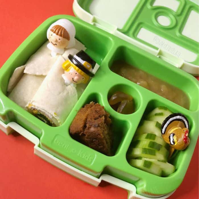 A bento lunchbox filled with wraps with pilgrim toothpicks, banana bread, cucumbers, gummies and applesauce
