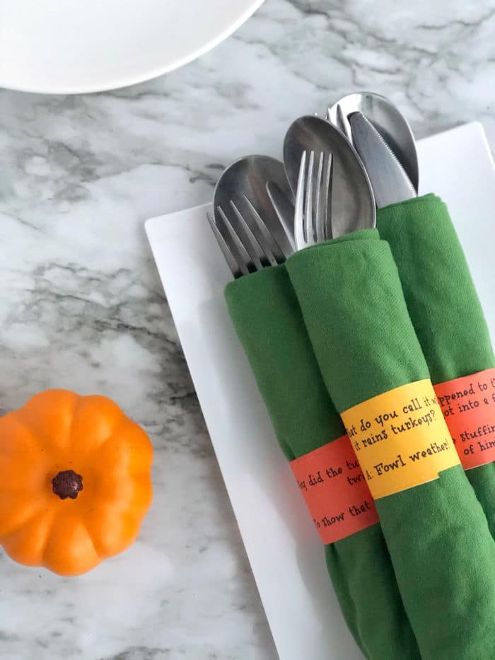 Silverware wrapped in napkins with jokes printed on the napkin rings, a decorative pumpkin next to them