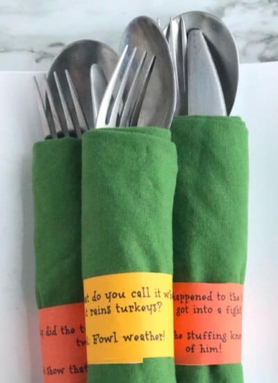 Three bundles of silverware wrapped in napkins are shown on a platter wrapped in silly Thanksgiving joke napkin rings.