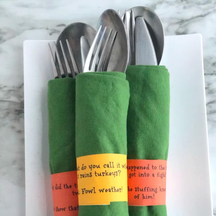 Three bundles of silverware wrapped in napkins are shown on a platter wrapped in silly Thanksgiving joke napkin rings.