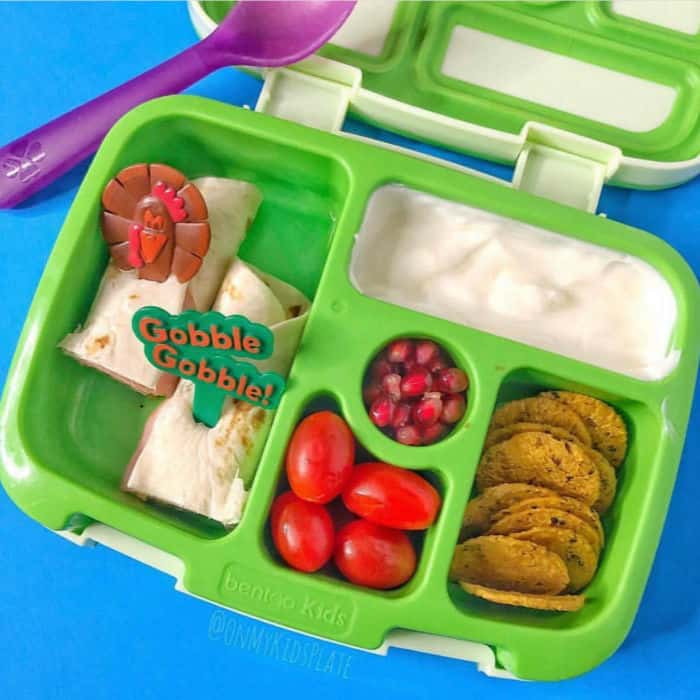 A bento lunchbox filled with tomatoes, pomegranate seeds, yogurt, wraps and sweet potato crackers