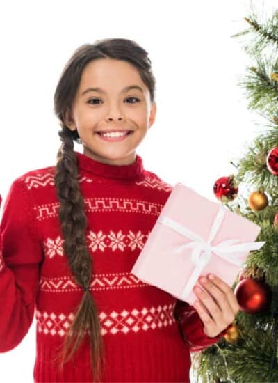 A little girl holding a present and standing next to a Christmas tree pointing to the side