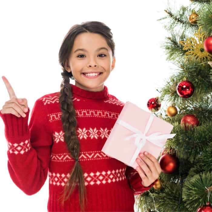 A little girl holding a present and standing next to a Christmas tree pointing to the side