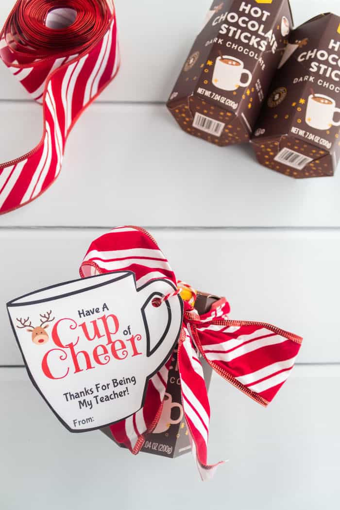A hot chocolate gift  being put together dressed up with a ribbon and a reindeer on a mug shaped gift tag that says \"cup of cheer, thanks for being my teacher\"