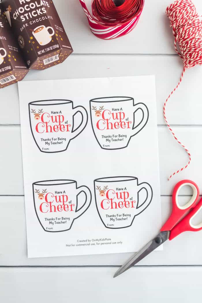 Mug shaped gift cards on a piece of paper that say \"Have a cup of cheer, thanks for being my teacher\" with a reindeer on them. Scissors and red string are also in the frame. 