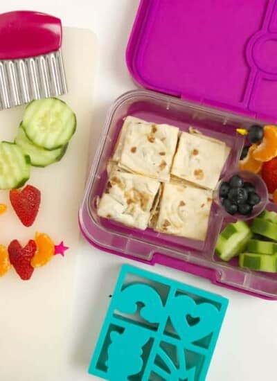 A kid's bento lunchbox full of quesadilla, cucumbers, blueberries, oranges and strawberries shaped like hearts