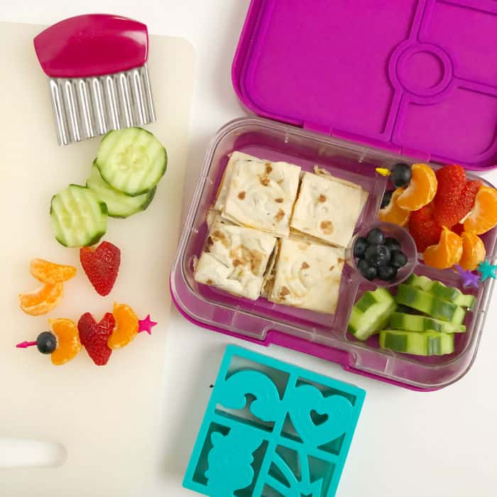 A kid's bento lunchbox full of quesadilla, cucumbers, blueberries, oranges and strawberries shaped like hearts