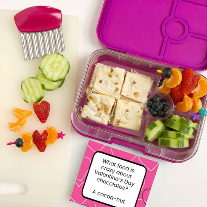 A kid\'s bento lunchbox full of quesadilla, cucumbers, blueberries, oranges and strawberries shaped like hearts with a lunchbox joke