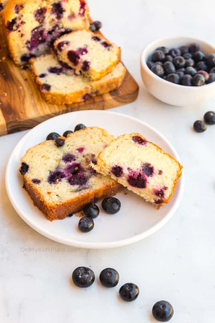 A piece of blueberry cake on a plate with more slices on a cutting board