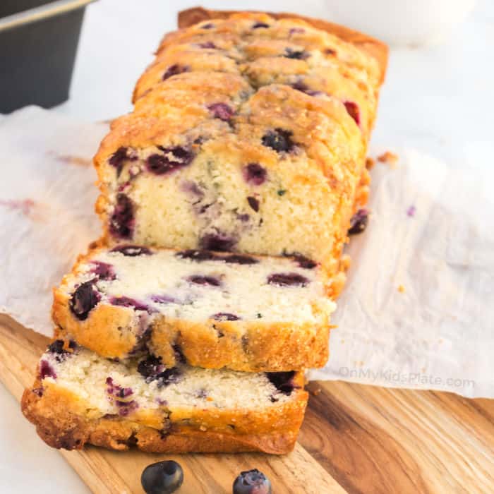 The end of a blueberry loaf, which sits sliced on a cutting board on top of a piece of parchment paper. A baking pan is in the background.